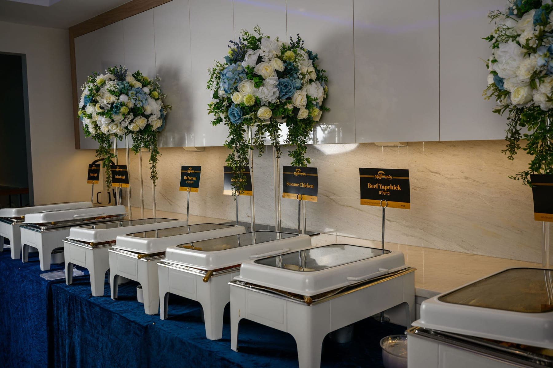 The buffet line laid for a vach nacht, twelve chafing dishes under tall floral arrangements, each tray behind its own Mashmanim name card